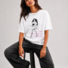Jeune femme brune posant en studio avec un t-shirt blanc large à motif graphique, un pantalon noir évasé et un sac à main noir à la main. Arrière-plan gris neutre.