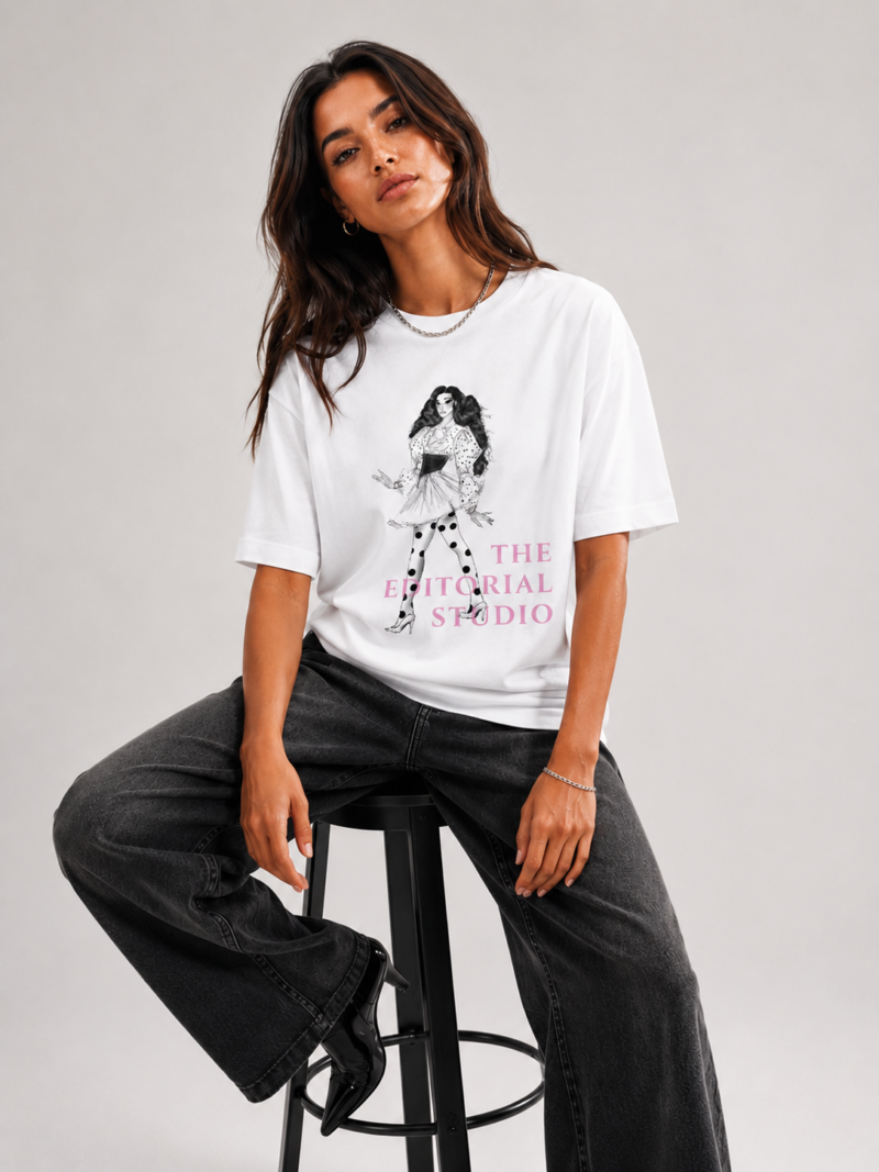 Jeune femme brune posant en studio avec un t-shirt blanc large à motif graphique, un pantalon noir évasé et un sac à main noir à la main. Arrière-plan gris neutre.
